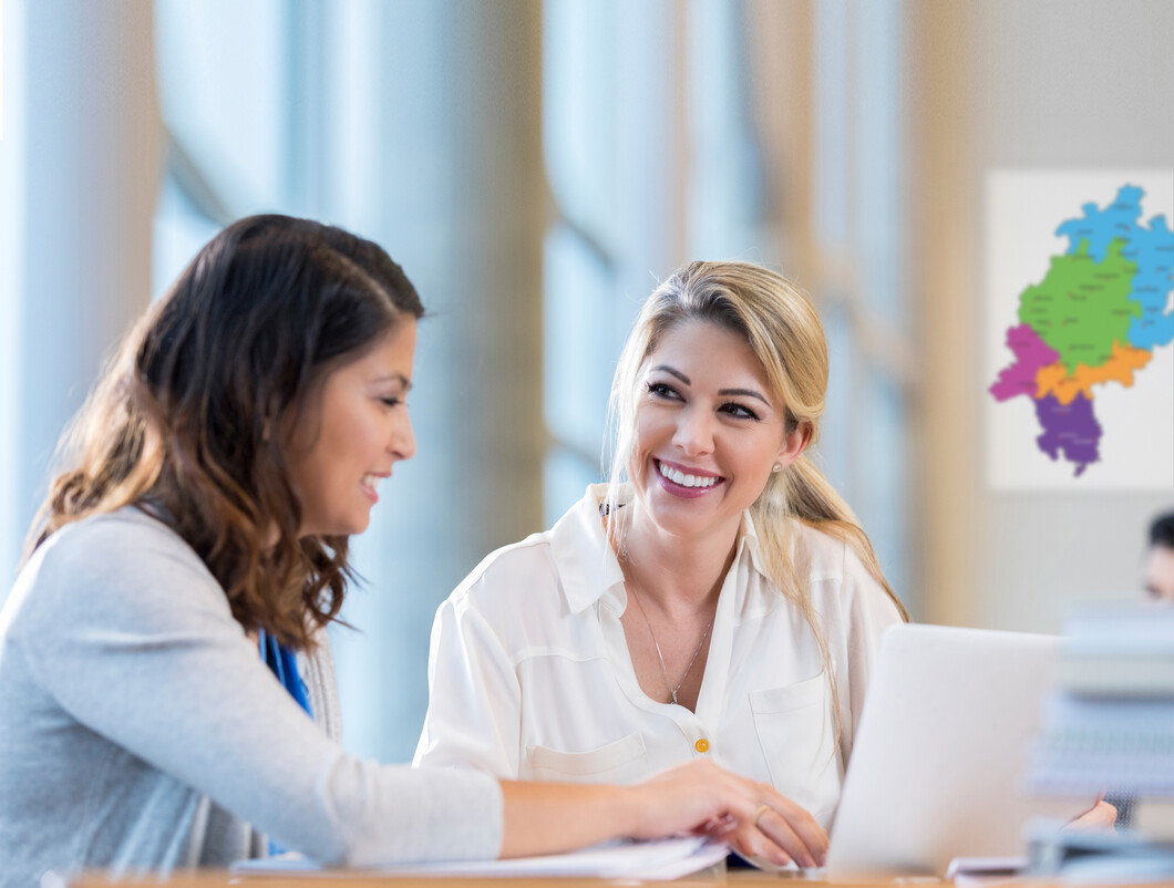 Zwei Frauen sitzen vor einem Schreibtisch mit einem Laptop. Die links sitzende Frau erklärt der rechts sitzenden etwas.  