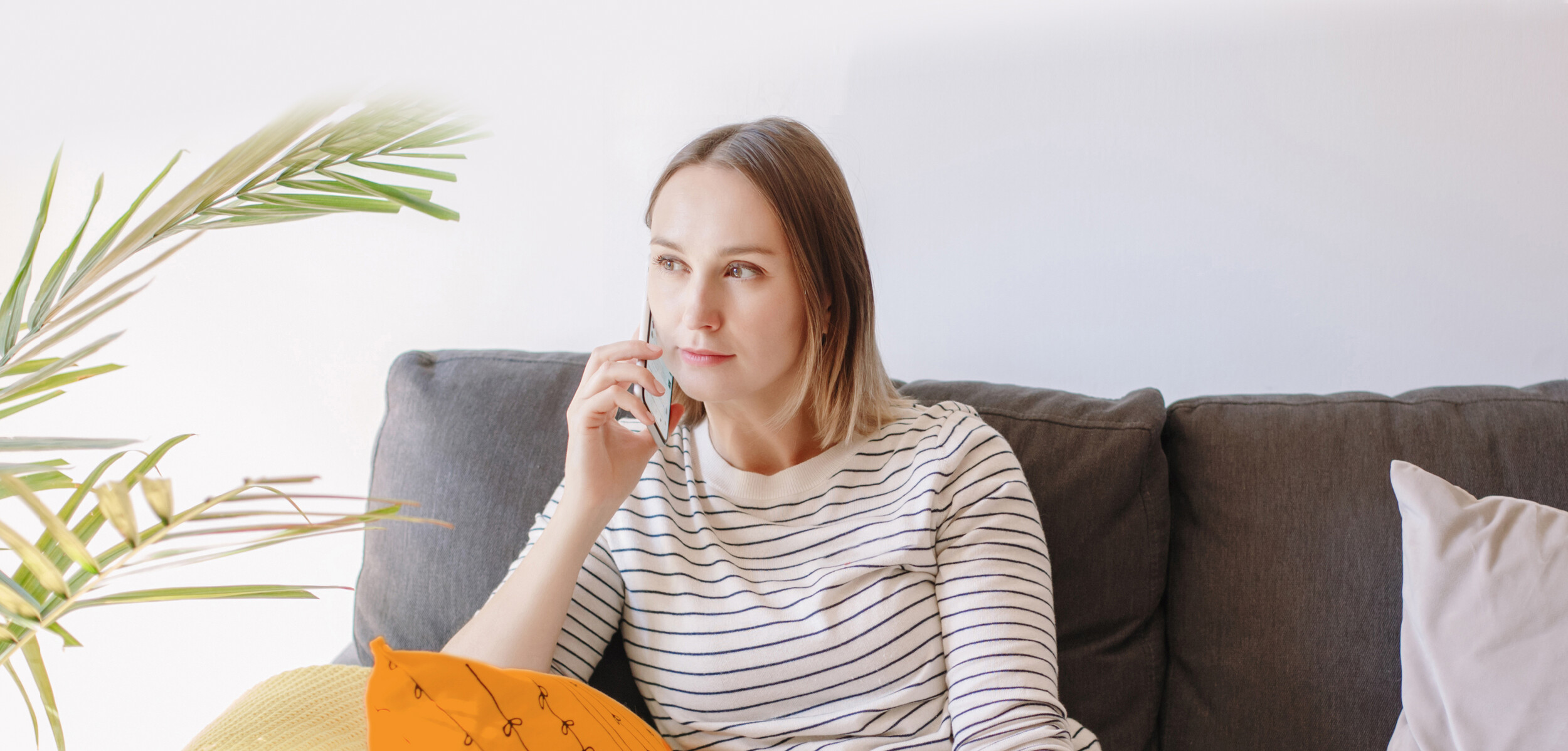 Junge Frau sitzt auf dem Sofa und telefoniert. Links von ihr steht eine Palme, im Vordergrund hält sie ein orangenes Kissen auf dem Schoß.