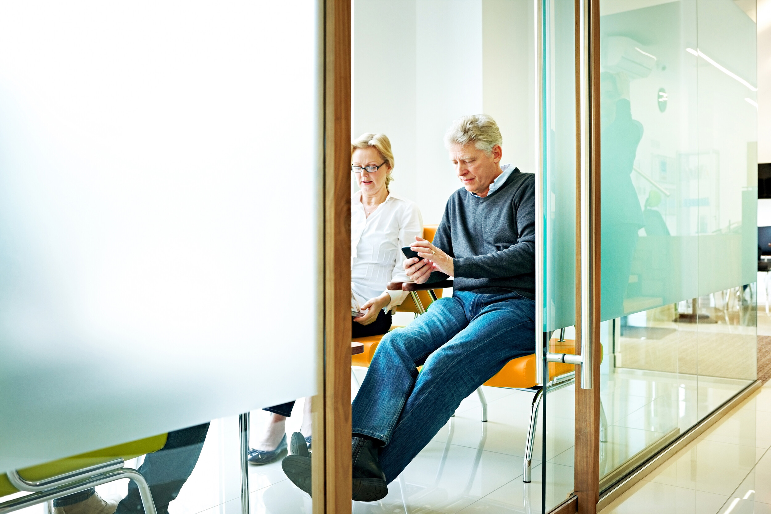 Zwei Patienten sitzen in einem Wartezimmer. Einer schaut auf sein Mobiltelefon. 