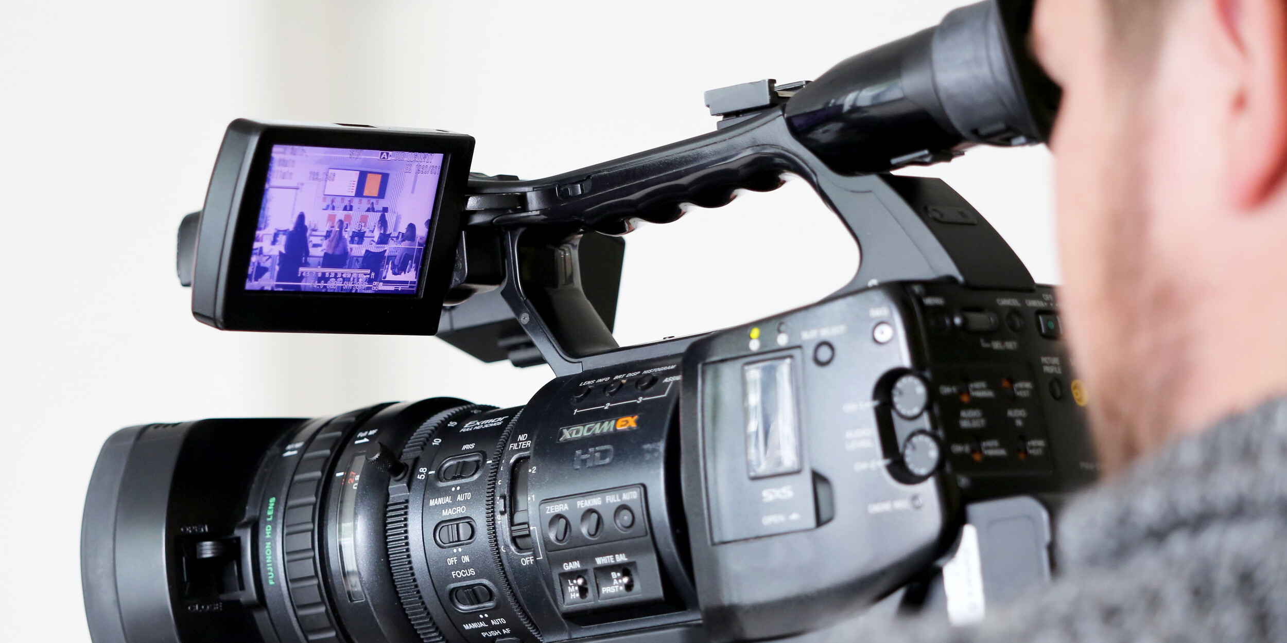 Ein Mann bedient eine professionelle Kamera. Im Bildschirm der Kamera ist eine Pressekonferenz zu sehen.