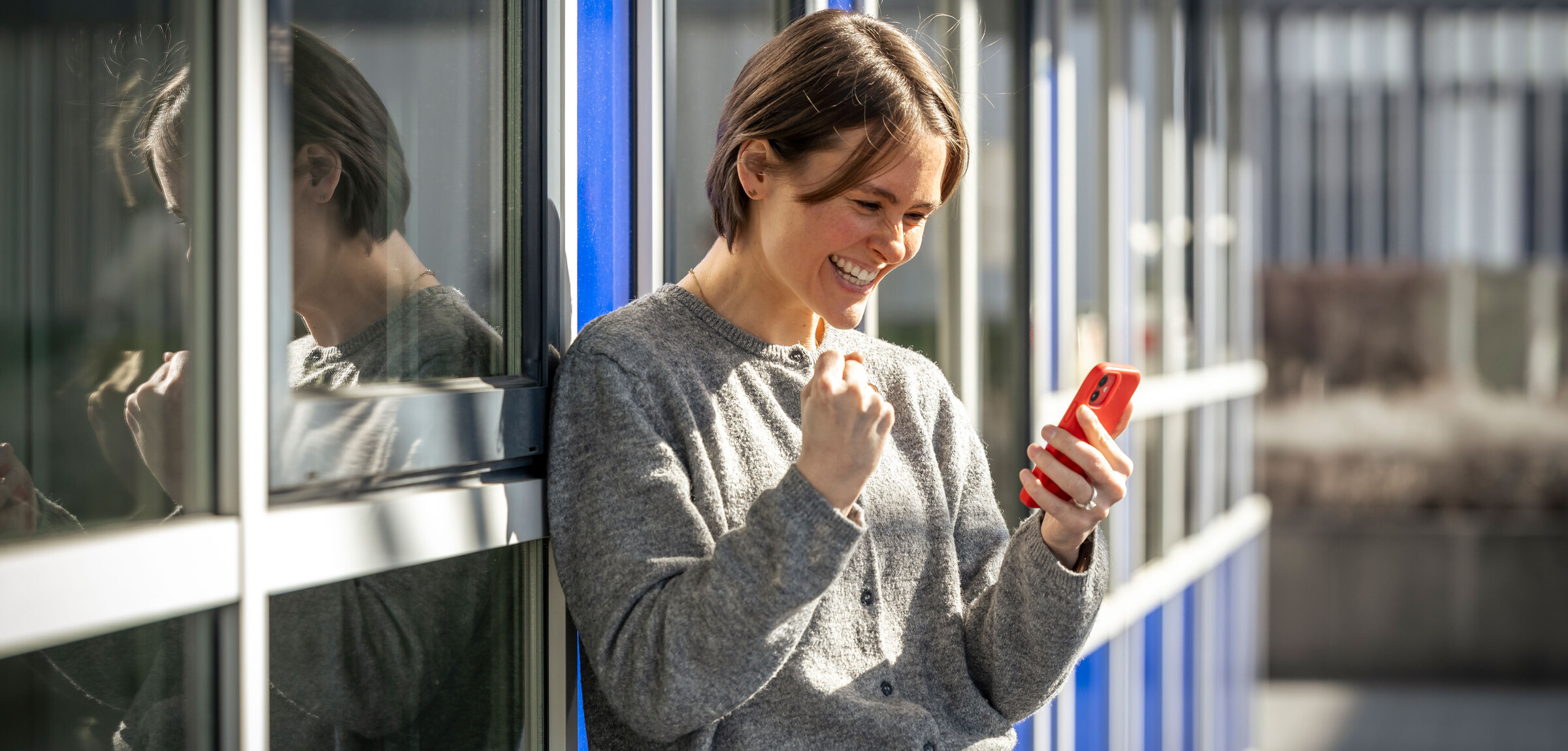 Eine Frau steht vor einem Bürogebäude und schaut erfreut auf ihr Handy.