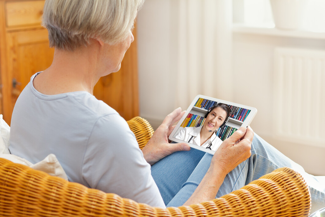 Eine ältere Frau sitzt zuhause in ihrem Sessel und hält ein Tablet in der Hand. Auf dem Bildschirm ist ihre Ärztin zu sehen.