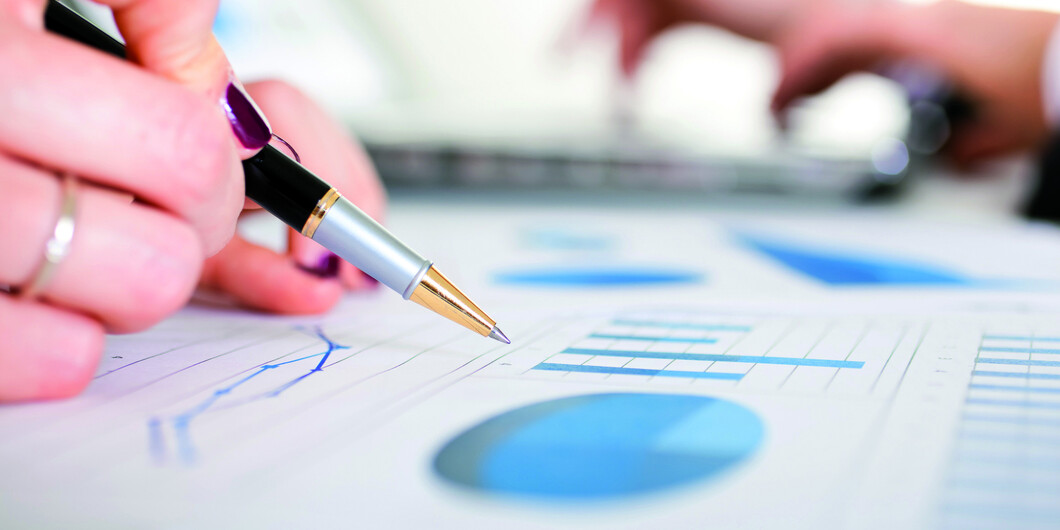 Hand mit Stift in den Fingern zeigt auf ein Blatt mit Auswertungen wie Grafen.