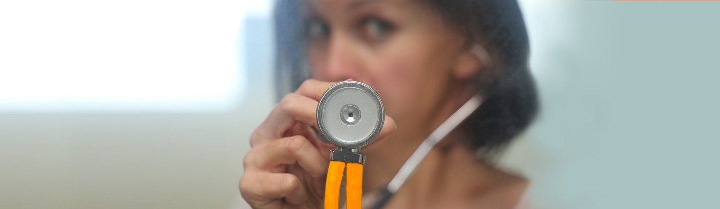 Eine Frau im Hintergrund hält den Kopf eine Stethoskops in Richtung Kamera