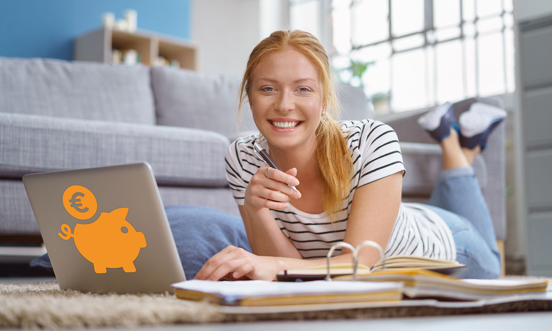 Eine Studentin auf dem Boden des Wohnzimmers, vor sich einen aufgeklappten Laptop, dessen Deckel ein orangener Sparschwein-Aufkleber ziert, und eine aufgeschlagenen Aktenordner