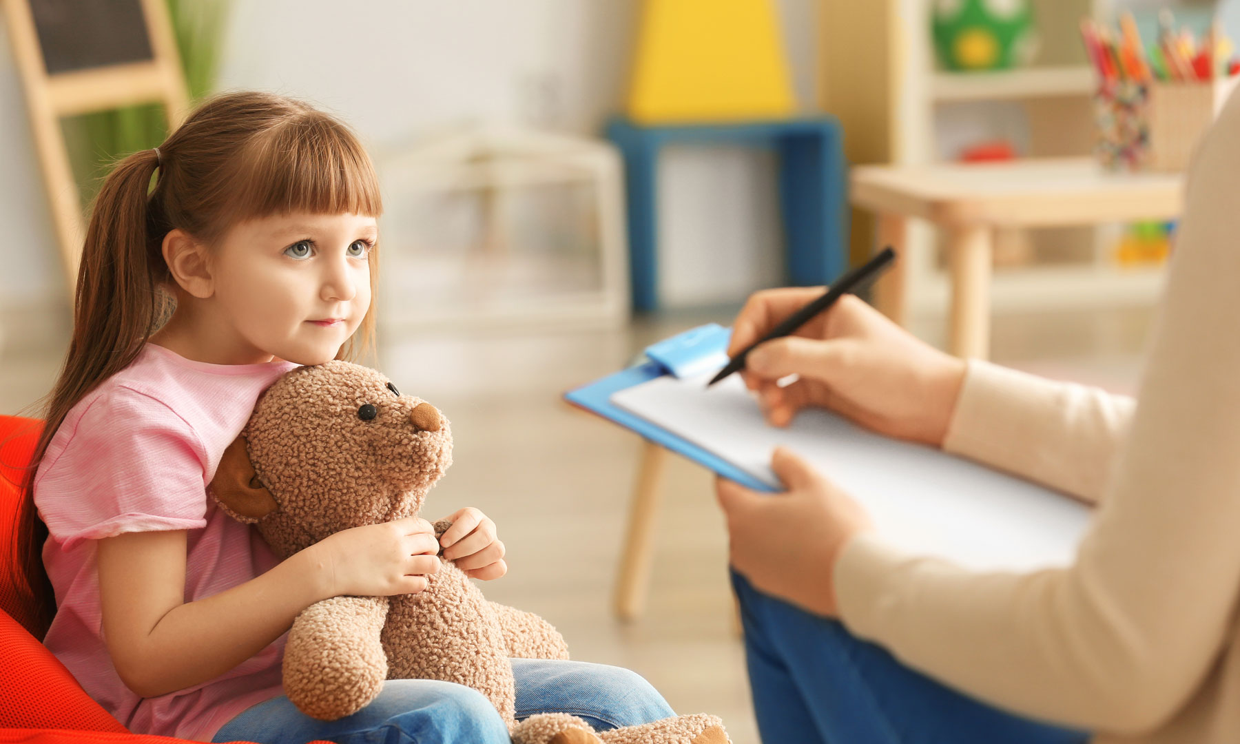 Ein Mädchen mit einem Teddy im Arm, gegenüber angeschnitten eine Therapeutin mit Klemmbrett und Stift n der Hand, die sich Notizen macht