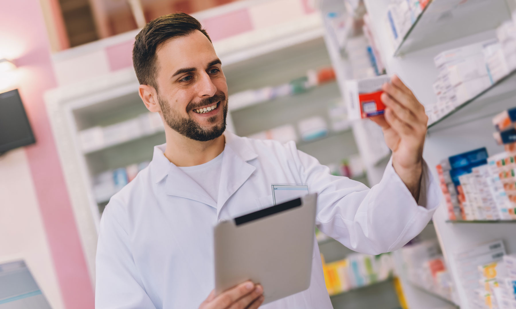 Mann mit weißem Kittel in Apotheke hält Schachtel mit Medikamenten.
