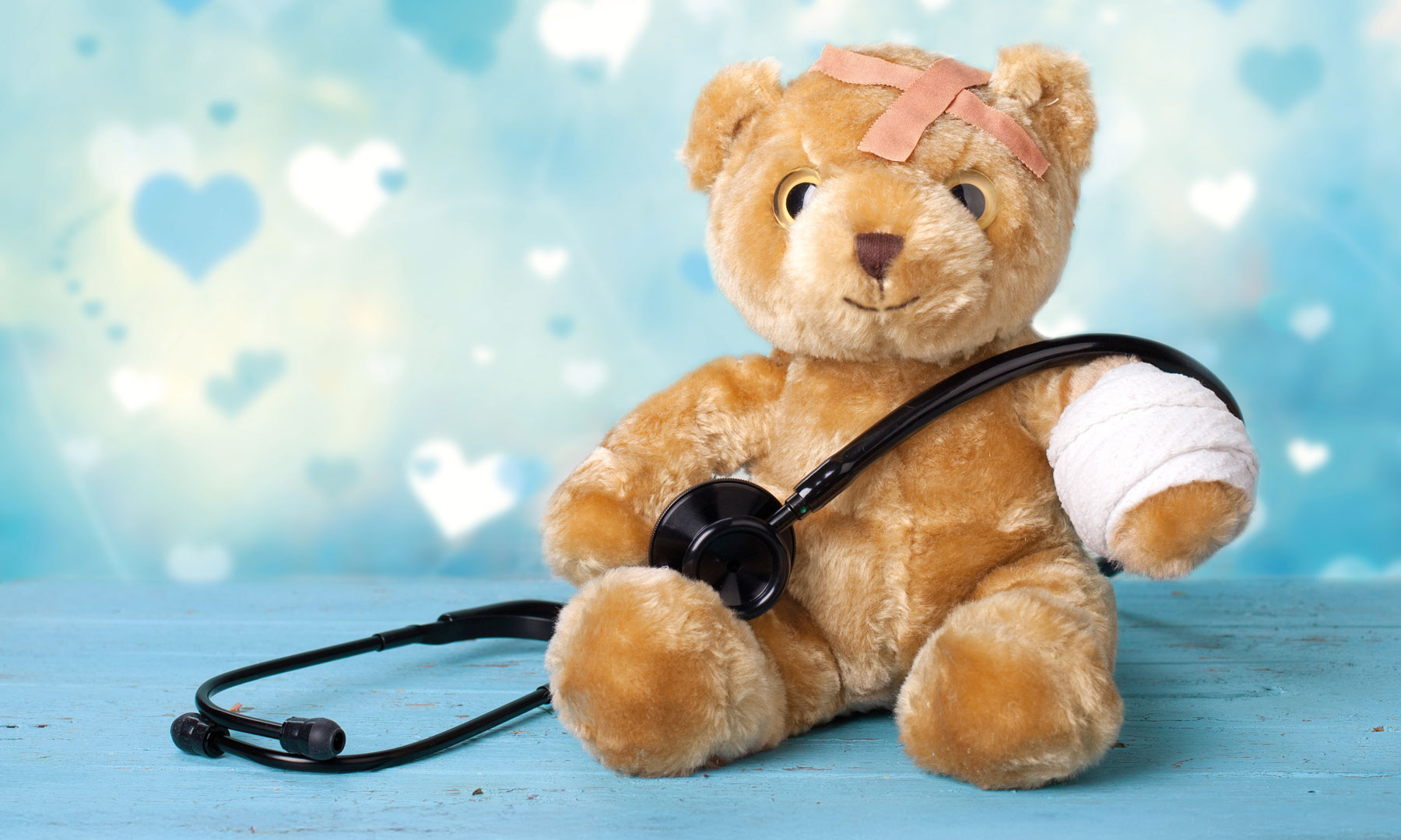 Ein Teddybär sitzt vor hellblauem Hintergrund. Er hat den rechten Arm verbunden und ein Stethoskop umgehängt.