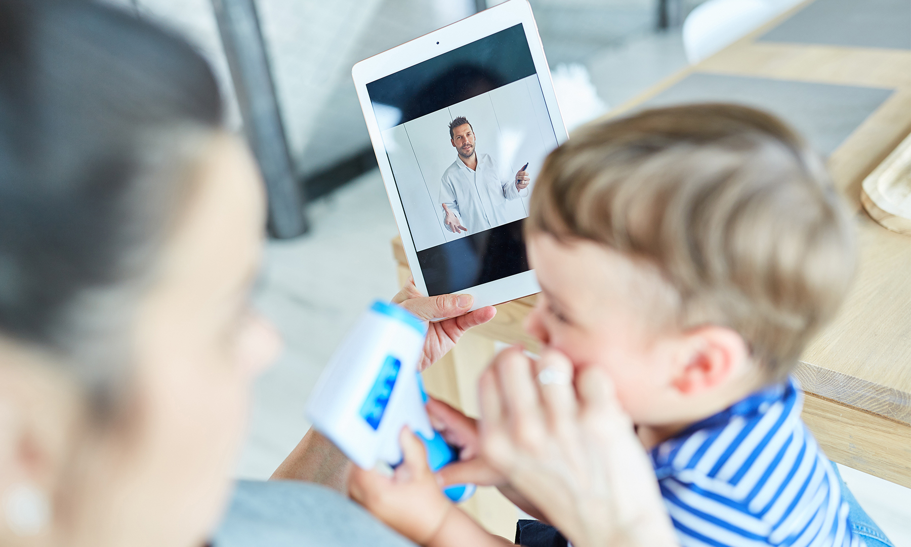 Mutter mit Kleinkind hat ein Tablet in der Hand auf dem ein Arzt in der Videosprechstunde zu sehen ist.